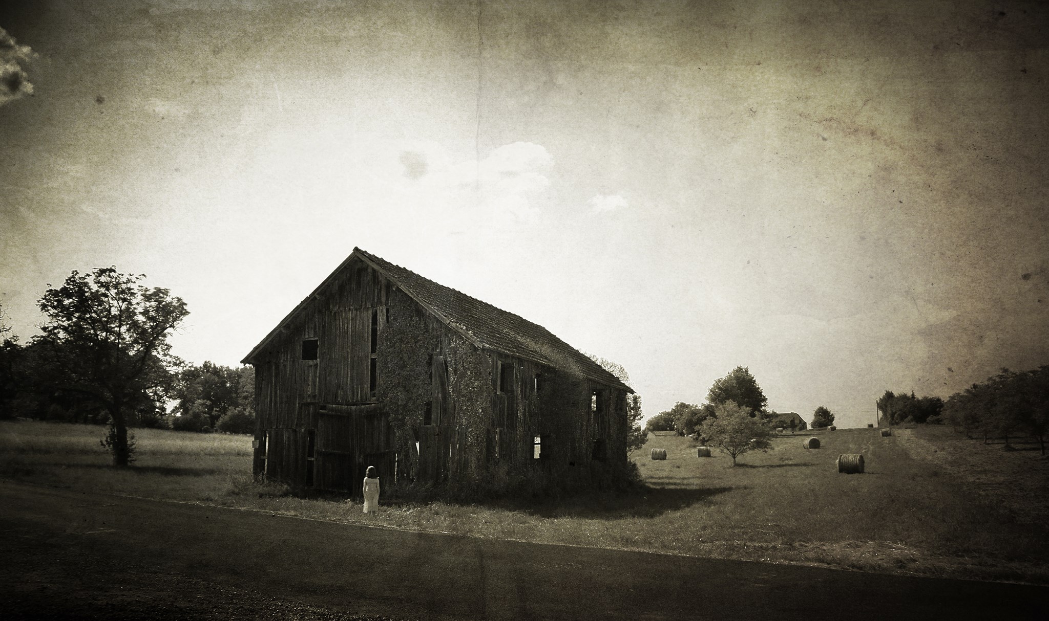 Photo sépia d'une vielle grange délabrée devant laquelle une enfant en chemise de nuit semble figée.